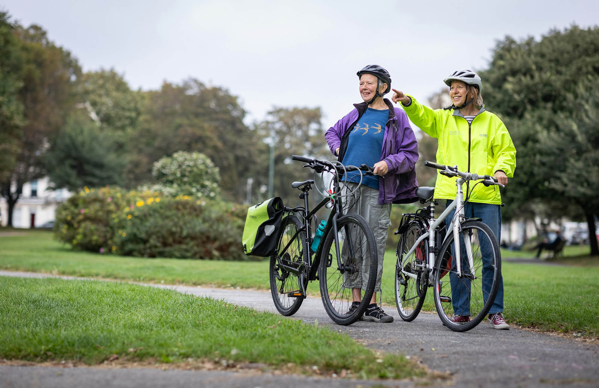 Två äldre personer står med sina cyklar i en park och pekar mot något