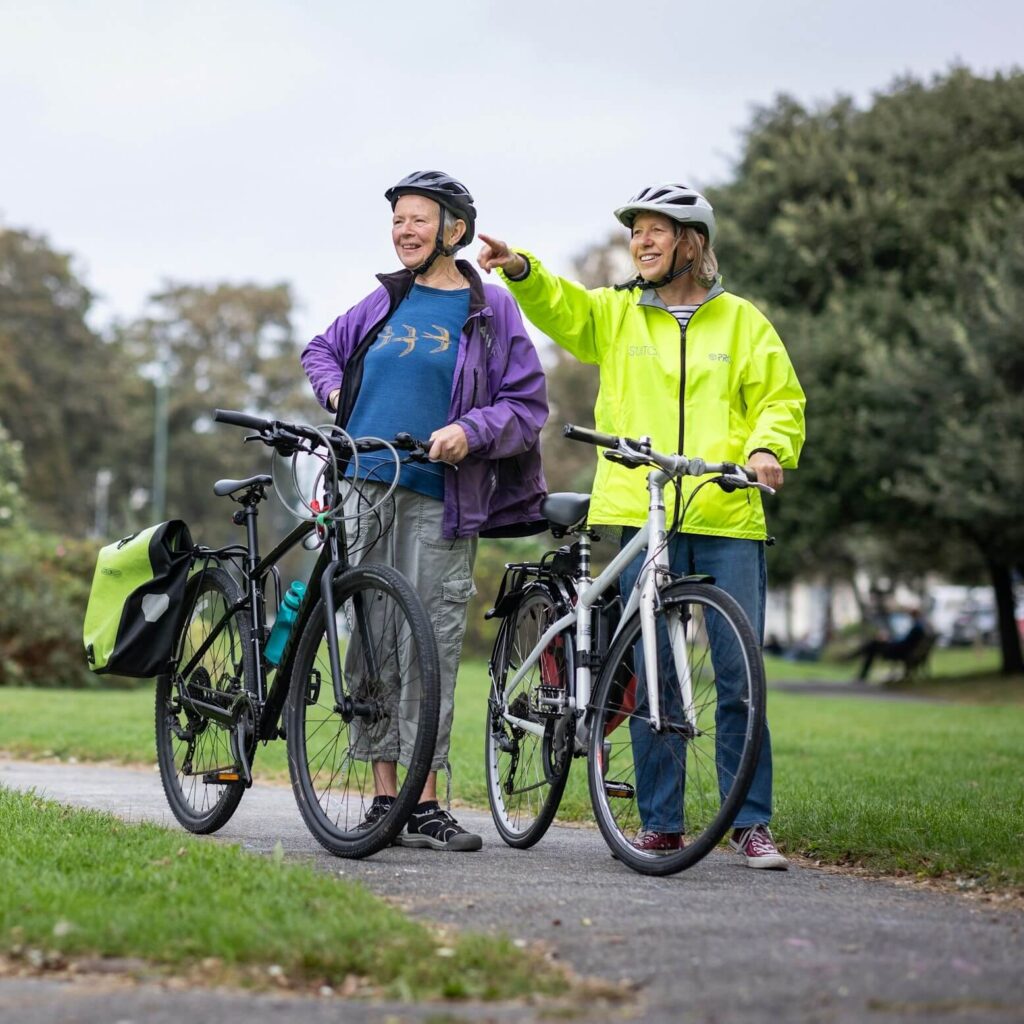 Två äldre personer står med sina cyklar i en park och pekar mot något
